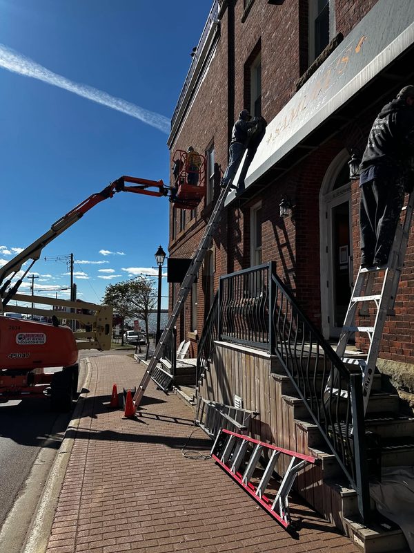Two workers, one on a boom lift and one on a ladder, stripping back the awning frame of a red brick commercial building