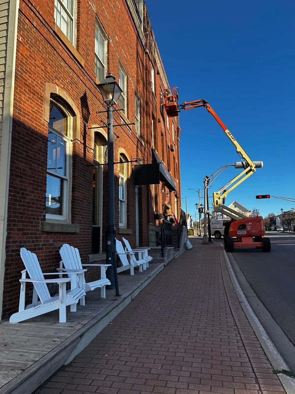 Looking down a brick sidewalk past white Adirondack chairs, with a boom lift reaching up to a red brick building further along