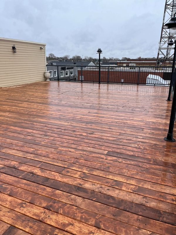 A rebuilt rooftop deck with freshly stained boards, a black metal railing, lamp standards and the harbour behind