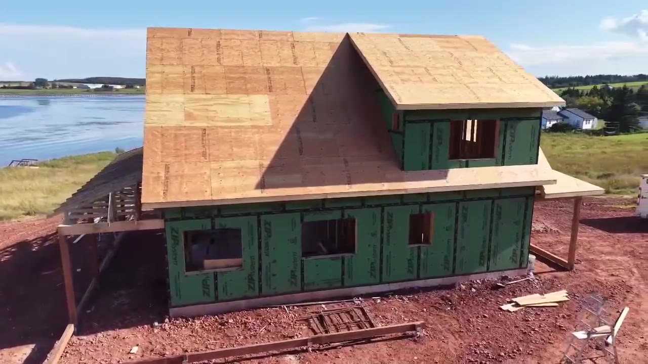 A new house under construction on a red-soil lot beside the water, green sheathing and fresh roof decking, filmed from the air