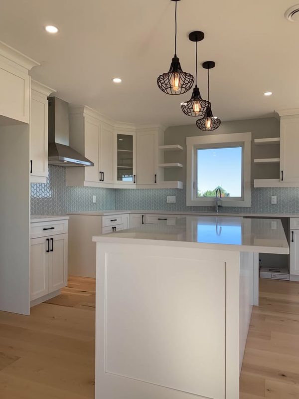 An all-white kitchen with black hardware, pale blue scalloped tile and three caged filament pendants over a quartz island